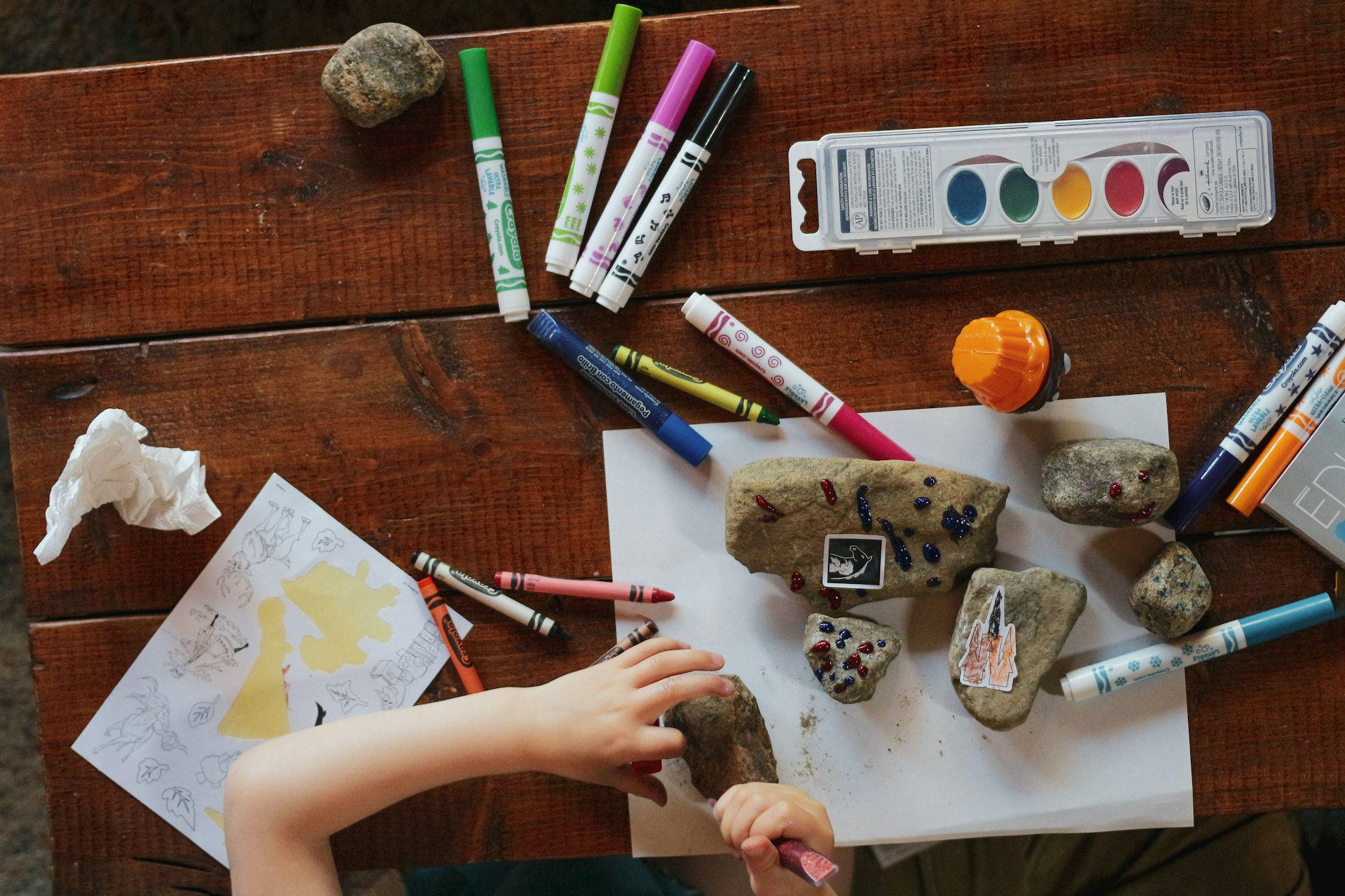 A child's hands working at an art table with crayons and paints